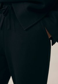 Čierne jogger nohavice z mäkkej textúrovanej tkaniny. Obsahujú pások so šnúrkou v páse, bočné vrecká a zúžené nohavice so šitými švami.