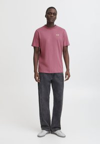 Rød T-shirt med korte ærmer og rund hals; har små guldtekster på brystet. Kombineret med løstsiddende mørkegrå jeans og hvide sneakers.