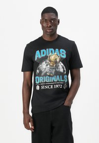 Fekete pamut póló, amelyen egy nagy grafikával ellátott trófea és az "ADIDAS ORIGINALS" felirat látható kék és fehér színben. Rövid ujjú, kerek nyakú.