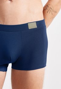 Marineblaue Boxershorts mit glatter Textur, ausgestattet mit einem SLOGGI-Logopatch am Bund und einem figurschmeichelnden Schnitt. Teilansicht eines Beins und des Oberkörpers.