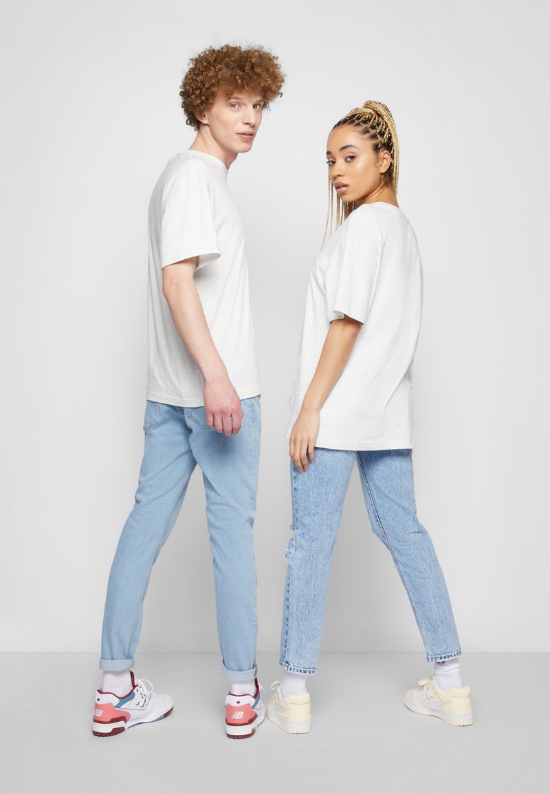 Due persone stanno schiena contro schiena indossando t-shirt grigio chiaro largo. L'uomo ha i capelli ricci ed indossa jeans blu; la donna ha i capelli intrecciati, jeans in denim con strappi e sneaker.