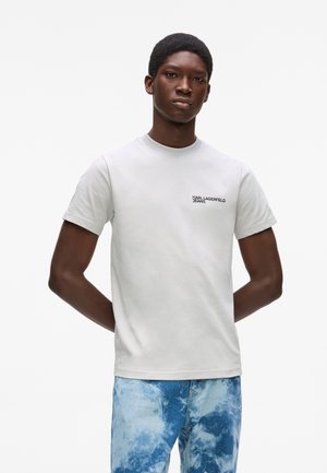 Jeune homme debout, les mains derrière le dos, portant un t-shirt blanc Karl Lagerfeld Jeans et un jean tie-dye bleu, sur un fond uni.