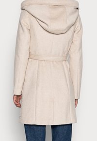 Abrigo de mezcla de lana beige con una gran capucha, cintura con cinturón y pliegues en la parte trasera. Presenta una textura suave y dos detalles de botones cerca del dobladillo.