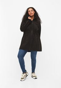 Zwarte, ritsjack met een trekkoord in de taille, voorzien van twee voorzakken. Gekoeld met blauwe jeans en meerkleurige sportschoenen.