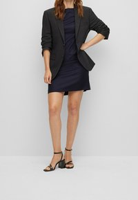 Blazer noir sur une robe bleu marine en tissu texturé. Le mannequin porte des talons noirs à lanières avec des talons carrés.