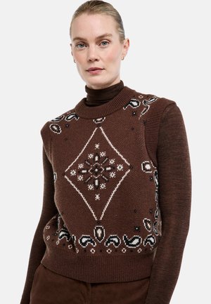 Femme portant un gilet en tricot sans manches à motif marron sur un col roulé marron foncé, avec des boucles d'oreilles et les cheveux tirés en arrière.