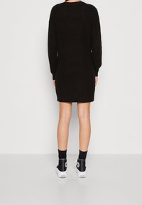 Robe en tricot noir avec des manches longues, coupe ample et texture côtelée. Portée avec des chaussettes noires et des baskets montantes blanches.