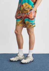 Pantaloncini colorati con motivo a scena desertica con vari animali. Indossati con scarpe da ginnastica beige e calzini bianchi, in piedi su un tappeto blu.