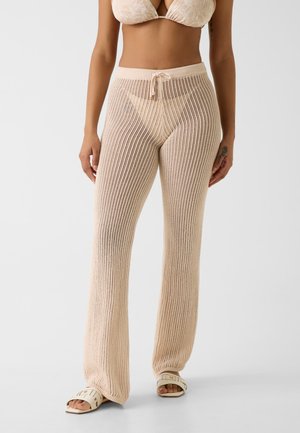 Femme portant un pantalon beige en maille à jambes larges avec une taille à cordon, associé à des sandales blanches à bouts ouverts et à un soutien-gorge clair assorti.