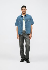 Mėlynas džinsinis marškinėlis trumpomis rankovėmis su dviem krūtinės kišenėmis, uždėtas ant balto marškinėlio su grafika, derinamas su pilkomis plačiomis kelnėmis ir juodais batais.