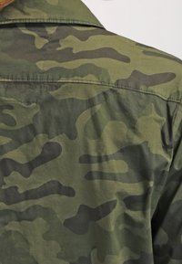 Tarnshirt in verschiedenen Grüntönen, mit einem strukturierten Stoff, einem Kragen und sichtbaren Nähten, das ein fließendes Muster zeigt.