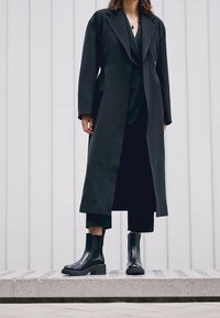 Czarny długi płaszcz z klapami z wycięciem, noszony na czarny strój. Modelka ma na sobie eleganckie czarne botki z grubą podeszwą, stoi na kamiennej platformie.