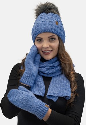 Blå strikket hue med pelskvast, matchende tørklæde og vanter. Varerne har et struktureret mønster og et brunt lædermærke.