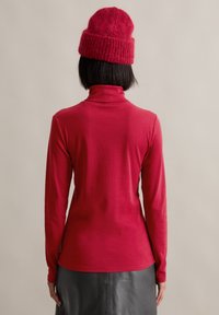 Red long-sleeve turtleneck top made of soft fabric. Features a fitted design and a high collar. Paired with a knitted red beanie.
