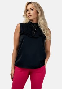 Czarna bezrękawnikowa bluzka z falbanianym kołnierzykiem, teksturowanymi detalami i zdobieniami przy szyi, zestawiona z różowymi dopasowanymi spodniami.