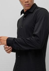 Zwarte button-up shirt in een gladde, glanzende stof met lange mouwen, puntige kraag en subtiele knoopdetails op de manchetten en de voorkant.