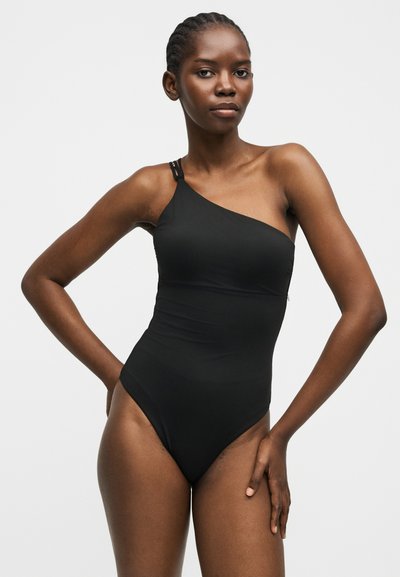 Γυναίκα με πλεξούδα που φοράει μαύρο, μονομάτικο μαγιό, στέκεται με το ένα χέρι στη μέση σε απλό γκρι φόντο.