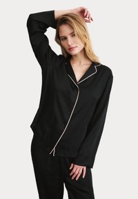 Svart satin pyjamas-set med långa ärmar, knapplås, rosa piping längs kragen och en avslappnad passform. Slät textur och lätt design.