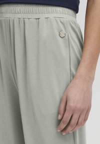 Ljusgrå byxor med en slät yta, utrustade med ett elastiskt midjeband och en dekorativ metallknapp på sidan.