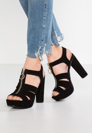 Czarne zamszowe szpilki na platformie z wyciętymi paskami i zamkiem błyskawicznym z boku, na tle znoszonych niebieskich dżinsów na białej podłodze.