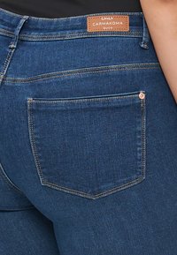 Dark blue denim jeans featuring a back pocket with yellow stitching and a copper-colored rivet. Leather brand patch at the waist.