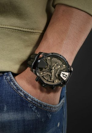Schwarze analoge Uhr mit transparentem Ziffernblatt, metallischen Akzenten und einem klobigen Design. Am Handgelenk getragen, kombiniert mit einem grünen Sweatshirt und Jeans.