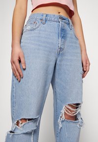 Jeans in denim azzurro chiaro con vestibilità rilassata, caratterizzati da due grandi buchi strappati sulla parte anteriore e un alto punto vita. Chiusura con semplice bottone di metallo.