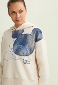 Felpa crema con un grande graphic di Topolino blu con contorni disegnati. Il materiale sembra soffice con una vestibilità comoda e una tasca frontale.