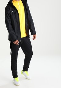 Black Nike jacket, yellow zip-up top, black pants with white accents, and lime green cleats. Features lightweight material and sporty design.