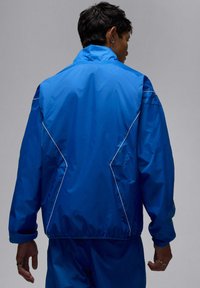 Blå vindjacka med hög krage, elastiska ärmslut och kontrasterande vita sömmar. Mjukt tyg med avslappnad passform och utan synliga logotyper.