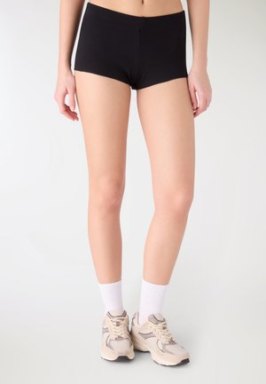 Donna che indossa pantaloncini neri, calze bianche a costine e scarpe da ginnastica beige, in piedi contro uno sfondo chiaro e uniforme.