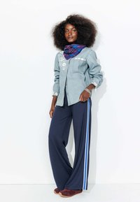 Chemise en denim avec broderies florales blanches, associée à un pantalon large bleu marine avec des rayures blanches sur les côtés et un foulard en tartan violet. Baskets bordeaux.