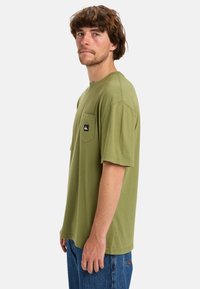 Olive green cotton t-shirt with short sleeves, crew neck, and left chest pocket; features a black brand patch. Casual fit and smooth texture.