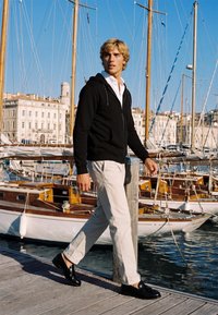 Jeune homme en sweat à capuche noir et pantalon blanc marchant sur un quai en bois près de voiliers dans une marina ensoleillée avec un ciel bleu clair.