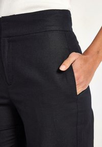 Pantalon noir avec une coupe droite, texture lisse, ceinture élastique et poches latérales. Le tissu a un léger éclat.