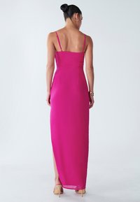 Duga, prianjajuća haljina od jarke ružičaste tkanine, s tankim trakama i bočnim prorezom, s glatkom teksturom i elegantnim dizajnom.