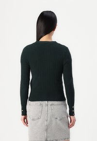 Dark green ribbed sweater with a round neckline and long sleeves, featuring gold button accents at the cuffs. Worn over a light gray denim skirt.