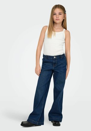 Ragazza giovane con lunghi capelli biondi indossa una canotta bianca senza maniche, jeans blu scuro a gamba larga, scarpe nere, in piedi davanti a uno sfondo chiaro e semplice.