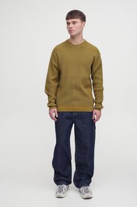 Sweter z fakturowanej dzianiny w oliwkowym kolorze z okrągłym dekoltem, w połączeniu z luźnymi, ciemnoniebieskimi dżinsami i jasnoszarymi sneakerami.
