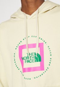 Felpa beige con il logo verde "The North Face" all'interno di un quadrato rosa, circondata da un testo circolare verde che legge "Non smettere mai di esplorare."
