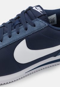 Gros plan sur une chaussure Nike bleu marine avec un logo swoosh blanc, des œillets en daim texturé et des lacets bleu foncé.