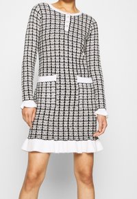 Abito a maglia a quadri bianchi e neri con maniche lunghe, scollo rotondo, pattina con bottoni, due tasche e orlo a balze bianche.