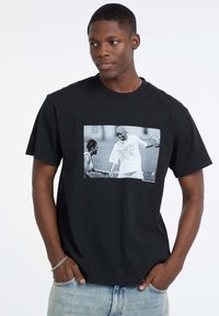 T-shirt en coton noir avec un imprimé graphique en niveaux de gris de deux joueurs de basket-ball. Encolure ronde, coupe décontractée et manches courtes.