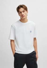 White cotton t-shirt with short sleeves, round neckline, and a small blue smiley face logo on the chest, paired with black cargo pants.