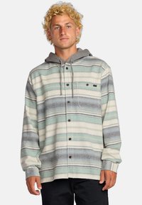 Camisa de franela a rayas en verde azulado, gris y blanco, con capucha gris, cierre frontal con botones y bolsillo en el pecho. Tejido suave con un corte relajado.