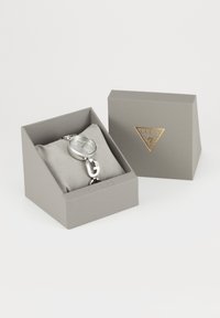 Silberne Guess-Armbanduhr mit rundem Zifferblatt und dekorativen Gliedern, präsentiert auf einem grauen Kissen in einer offenen grauen Guess-Box.