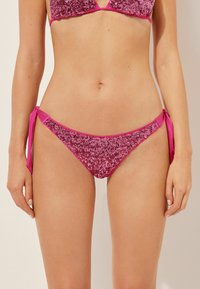 Rosa Pailletten-Bikini-Oberteil mit seitlichen Bindebändern. Glänzende Textur und kontrastierende einfarbige Akzente. Verstellbare Schleifen an den Hüften.