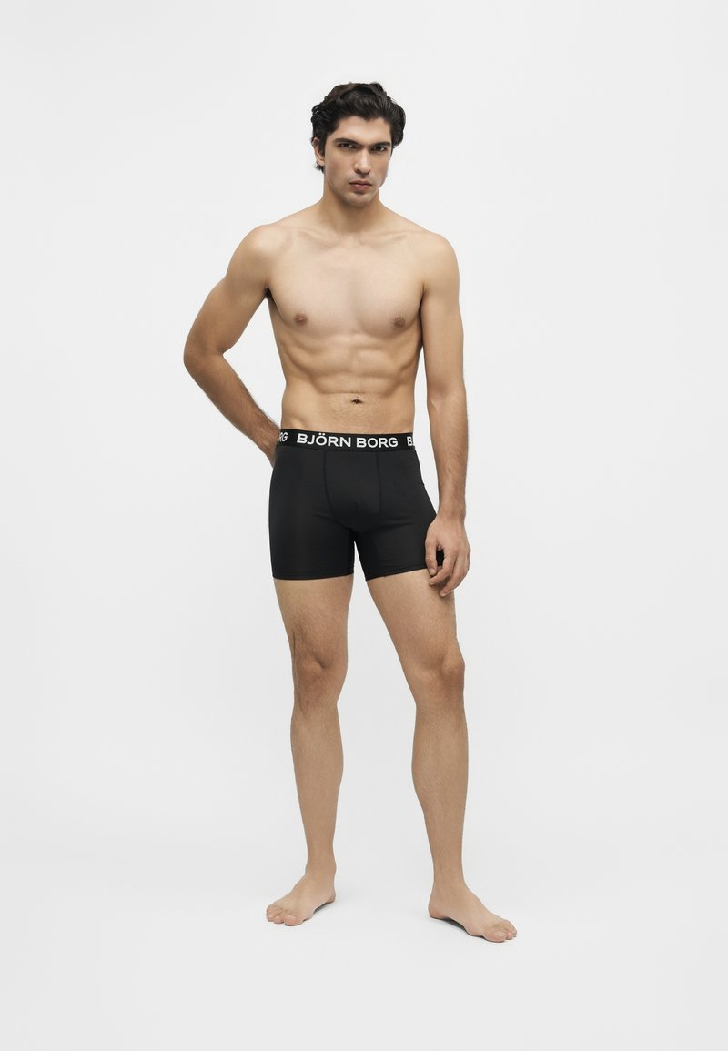 Homme torse nu, debout pieds nus, portant un boxer noir Björn Borg sur un fond clair uni.