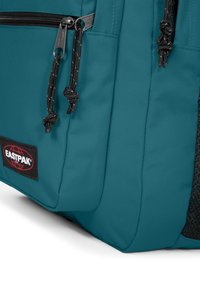 Tyrkysový látkový batoh s hladkou texturou, vybavený černými zipovými taháčky, jemným šitím a síťovanou boční kapsou. Zobrazeno logo Eastpak.