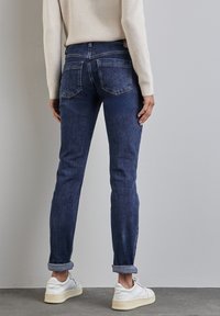 Jean en denim bleu marine avec une coupe ajustée, ourlets retroussés et deux poches arrière, associés à des baskets blanches sur un sol gris texturé.